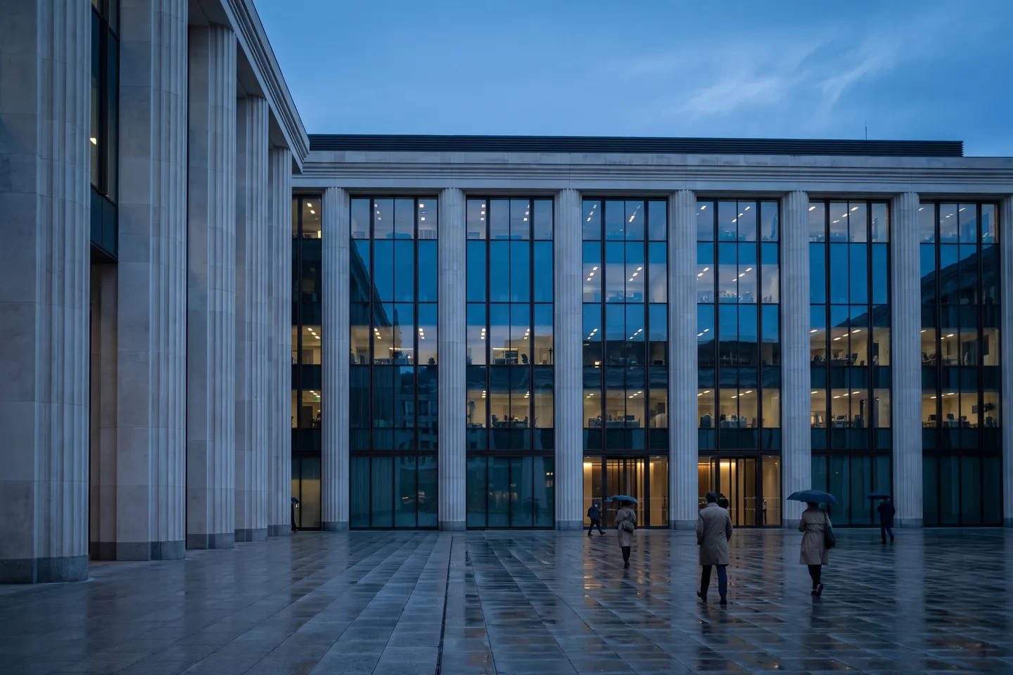 A columned stone and glass institutional building at blue hour, seen from a wet plaza